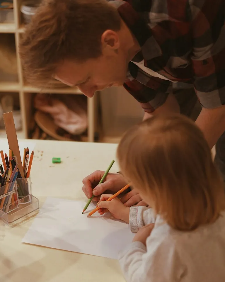 Un adulte accompagne un enfant qui dessine à une table.
